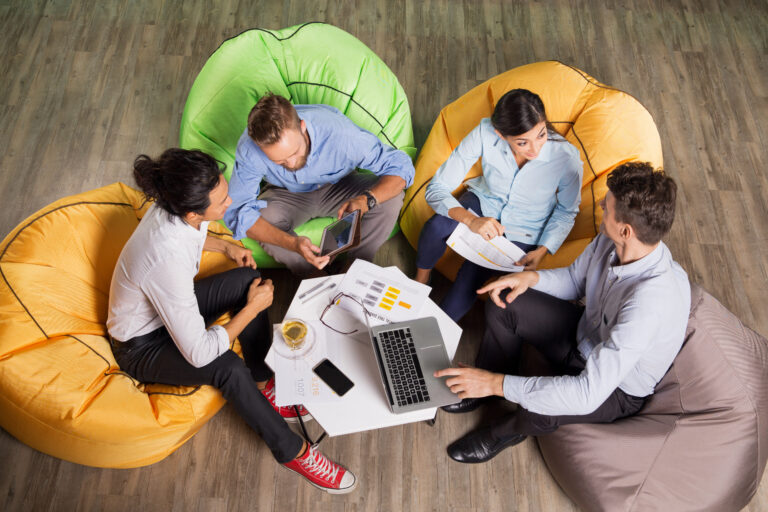 Four active young people sitting on beanbag chairs around small table in cafe, working and discussing ideas. High angle view.