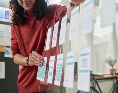 Young adult Caucasian man arranging mobile app interface mockups on transparent board, standing in modern office, focusing on user experience design process for digital project