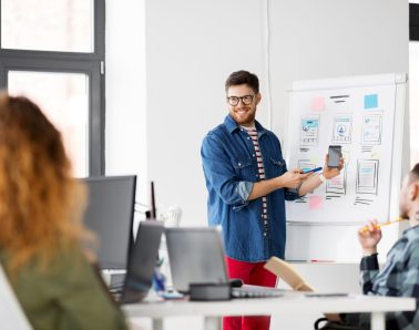business, technology and people concept - man showing smartphone user interface design to creative team at office presentation